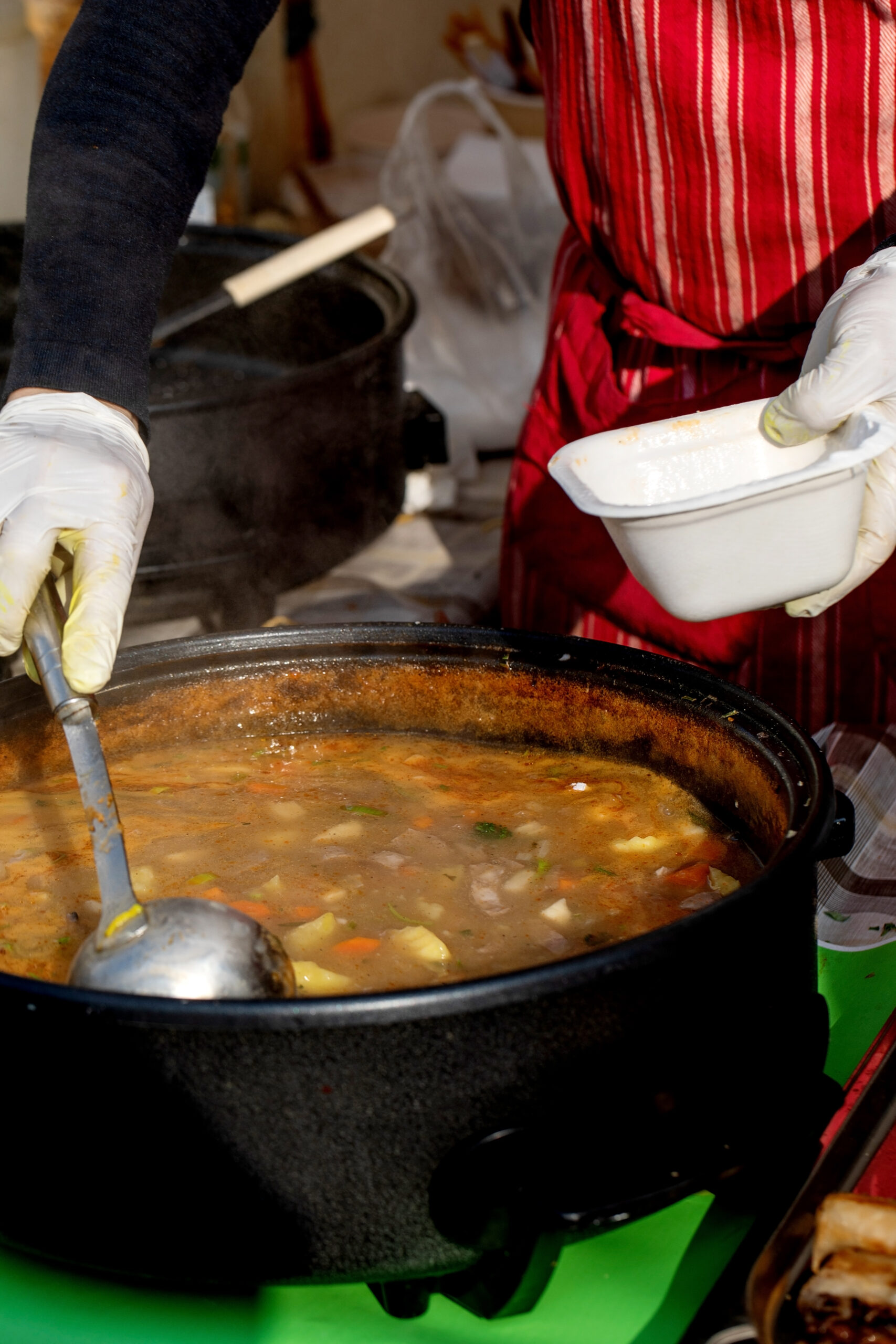 European street food festival. Vendor wearing disposable gloves serving traditional potato soup from large pot into takeaway bowl. Authentic homemade cuisine prepared fresh outdoor market setting